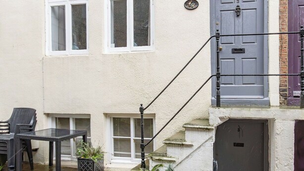 Ein zweistöckiges Gebäude mit weißer Fassade, einer grauen Tür und einer kleinen Außenbereich-Sitzgelegenheit mit Tisch und Stühlen.