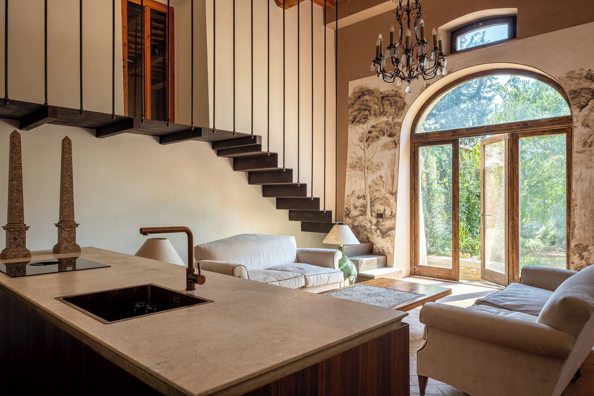 A modern kitchen with a built-in sink, a staircase with dark railings, and a large arched window with a view of greenery.