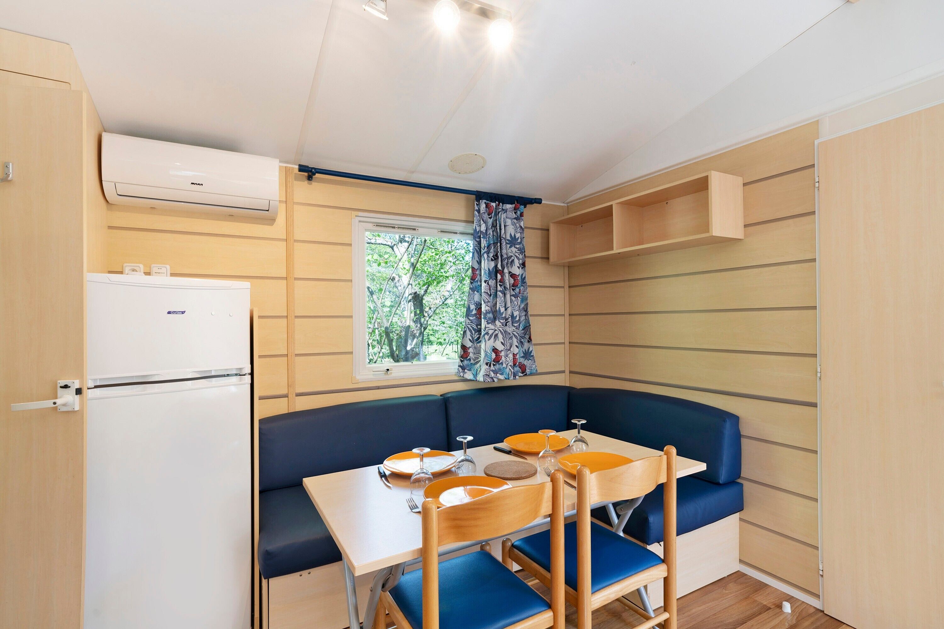 Une petite cuisine équipée comprenant un réfrigérateur, une fenêtre avec des rideaux, une table à manger avec des chaises et un banc intégré.