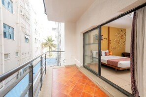 Habitación de hotel con cama, puerta de vidrio, balcón con barandilla y vista a edificios.