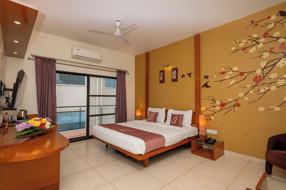 Ein Hotelzimmer mit Bett, Schreibtisch mit Blumenarrangement, Schreibtischlampe, Wandklimaanlage und einem mit Vorhängen versehenen Fenster.