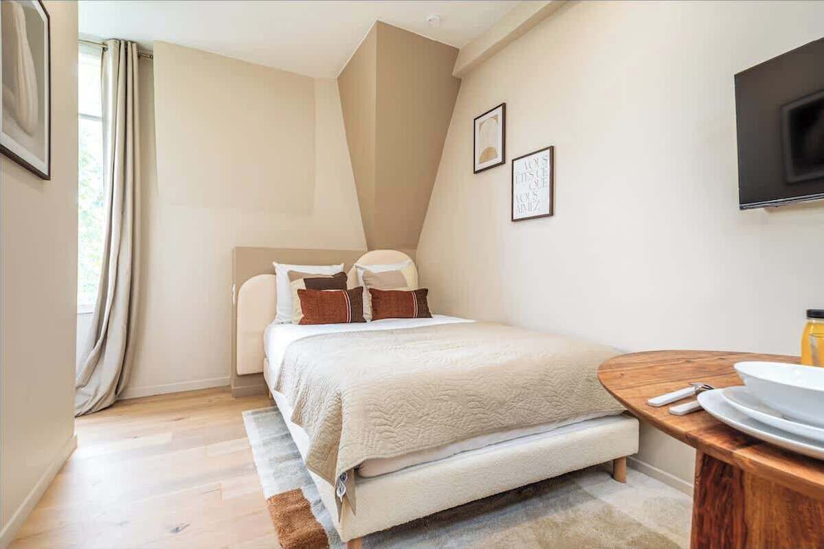 Een slaapkamer met een bed, een nachtkastje met een kom en een fles, een aan de muur bevestigde televisie en een raam met gordijnen.