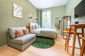 A compact room with a bed, a sofa, a dining table with chairs, a green rug, and a window with curtains. - Bright, air-conditioned studio for 2 people (Paris)