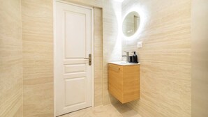 A bathroom with a white door, a wooden vanity with a mirror, and a light fixture on the wall. - Bright, air-conditioned studio for 2 people (Paris)