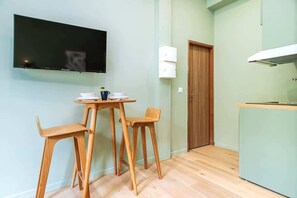 A small kitchenette with a TV mounted on the wall, a wooden table with chairs, and a door with a light switch. - Bright, air-conditioned studio for 2 people (Paris)