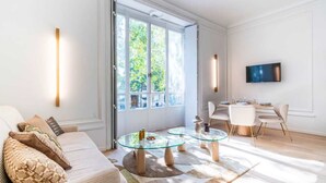 Ein modernes Wohnzimmer mit Glastisch, einer Couch mit gemusterten Kissen, einem Essbereich inklusive Fernseher und großen Fenstern.