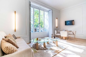 A modern living room with a glass coffee table, a sofa with patterned cushions, a dining area with a TV, and large windows. - Elegant 2-room air-conditioned apartment - Paris 17 (Paris)