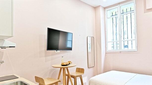 A hotel room with a bed, a TV, a small table with chairs, a window with bars, and a sink. - Studio climatisé, calme & tout équipé – Paris 17 (Paris)