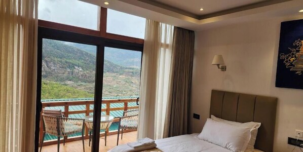 A hotel room with a bed, a view of a mountainous landscape through large windows, a balcony with a table and chairs, and a wall-mounted lamp. - Nangter Retreat (Paro)