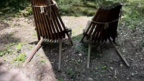 Deux chaises en bois rustiques, attachées à une branche d’arbre à l’aide de cordes, sont placées dans une zone boisée, près d’une étendue d’eau.