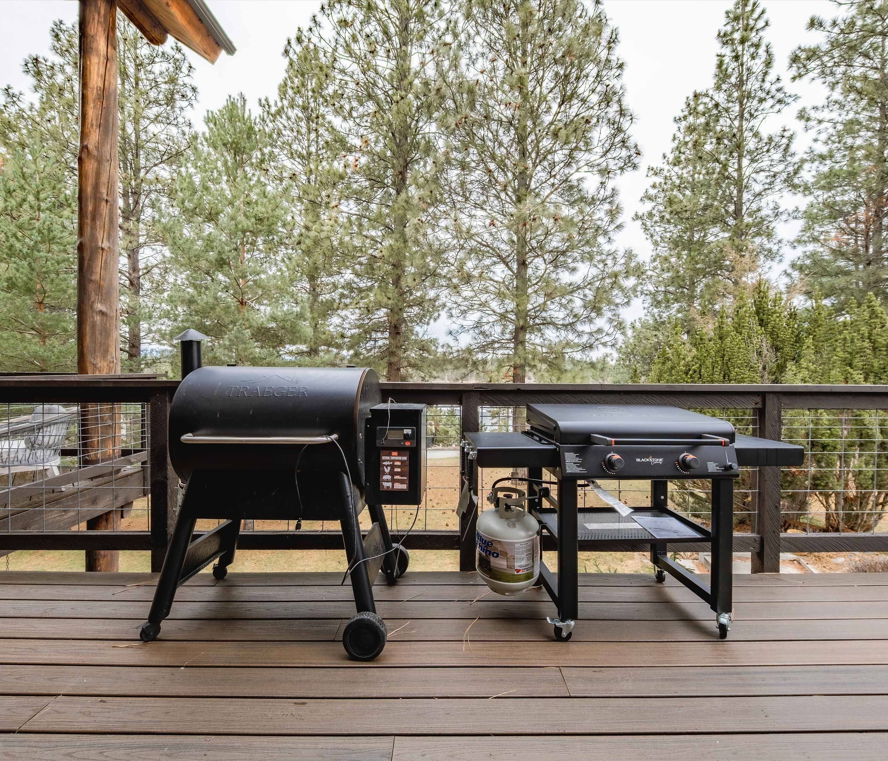 Un barbecue et un chariot pour barbecue sur une terrasse en bois, avec vue sur de grands pins.