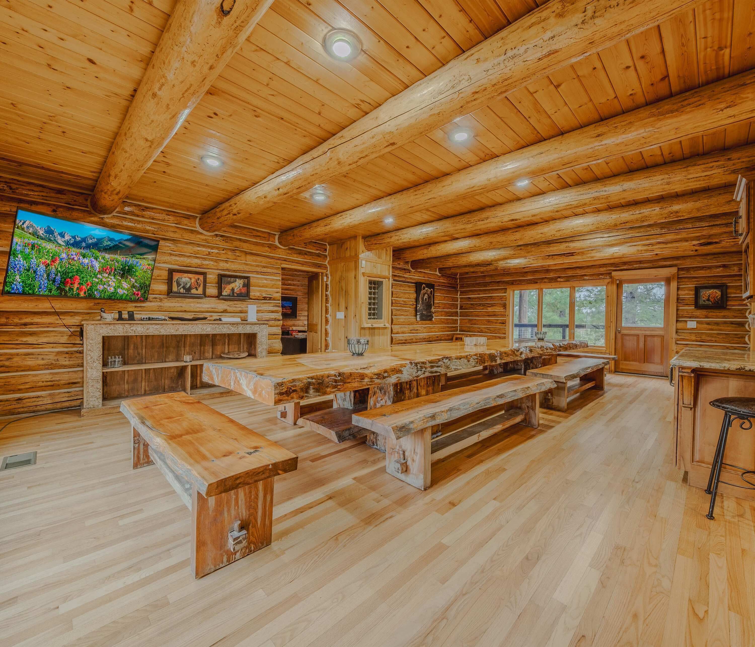 A rustic wooden cabin interior with a long communal dining table, benches, and a television mounted on the wall.