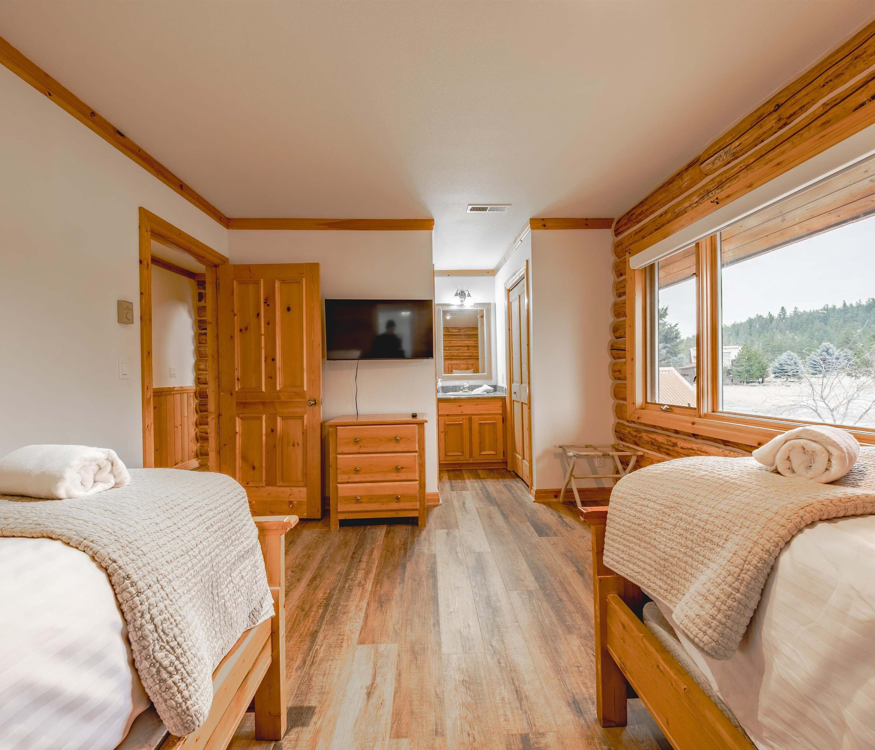 A cozy cabin room with two beds, wooden walls, a TV, and a window with a view of trees.