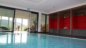 An indoor swimming pool with clear water, a glass door leading outside, and a red seating area with a plant. - Sontheims Naturhotel & Spa GmbH&Co.KG (Maierhöfen)