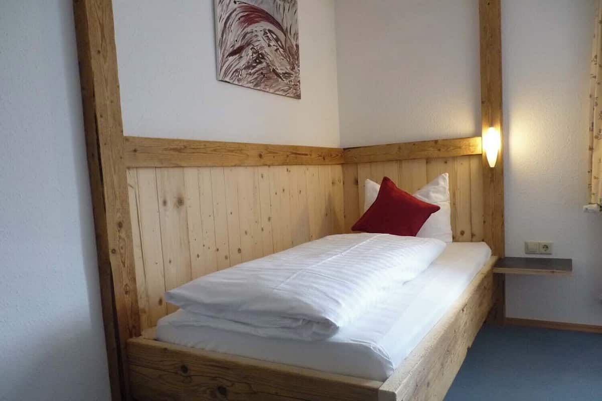 Ein Holzbett mit weißer Bettwäsche und einem roten Kopfkissen in einem Raum mit einem hölzernen Kopfteil und einem kleinen Regal.