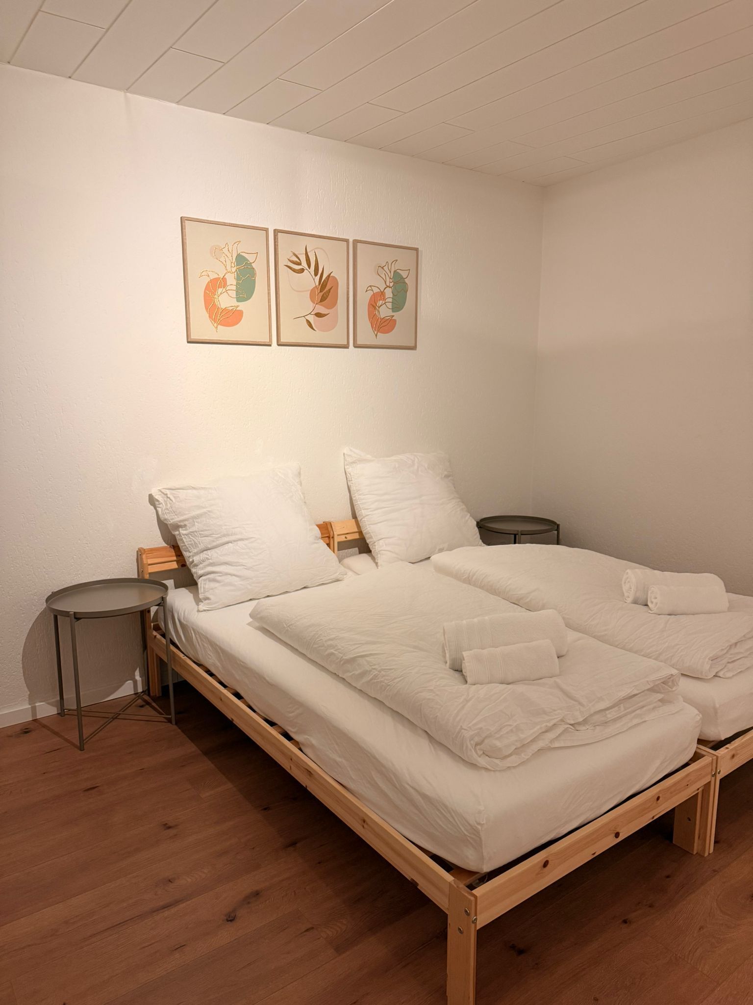 Una cama individual con estructura de madera, ropa de cama blanca y dos mesitas de noche. Hay tres cuadros enmarcados en la pared sobre la cama.