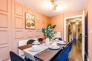 Un coin repas avec une longue table dressée pour quatre personnes, des assiettes blanches, des verres et un vase rempli de fleurs.