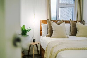 A neatly made bed with multiple pillows, a bedside table with a lamp and a small plant, and a window with a view of a brick building.