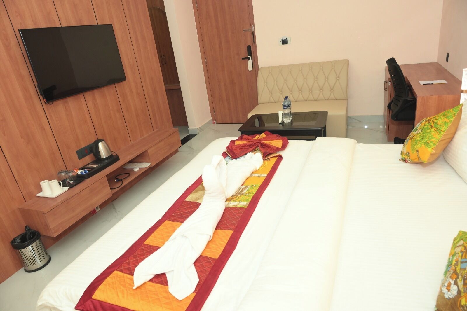 A hotel room with a bed, a TV mounted on the wall, a wooden cabinet, a small table, and a bench.