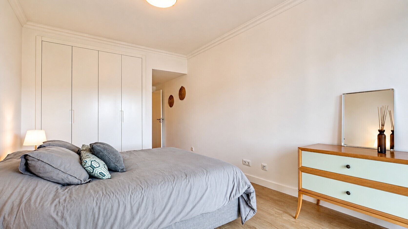 Een slaapkamer met een bed, een linnenkast, een spiegel en een deur die naar een andere kamer leidt.