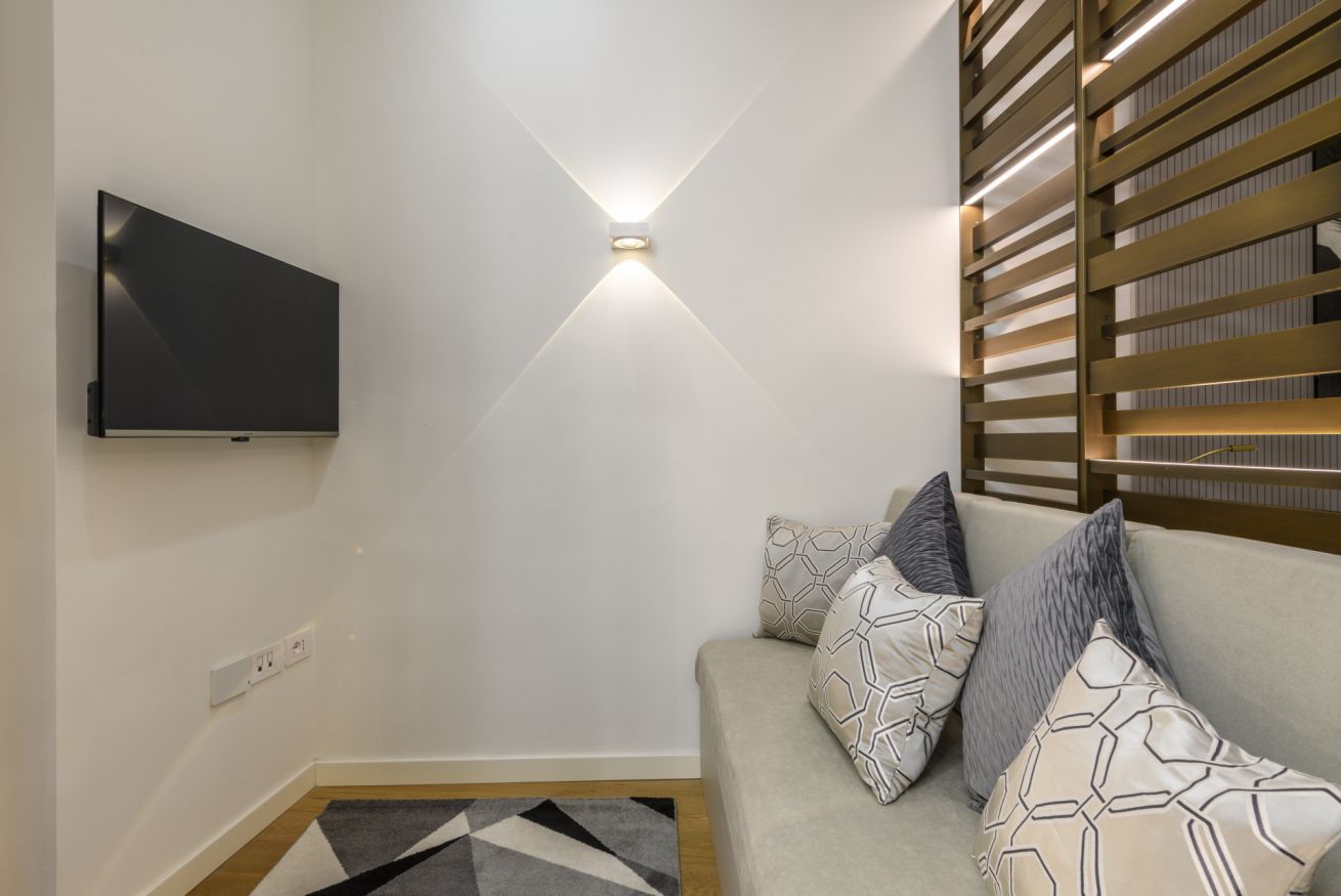 Un salon moderne doté d’un téléviseur à écran plat fixé au mur, d’un luminaire, d’un canapé avec des coussins à motifs et d’une cloison en lattes de bois.