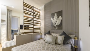 A bedroom with a bed, a window with blinds, a painting on the wall, and a bedside table with a lamp. - Gian Giacomo Mora, 13 - Refined studio apartment downtown (Milan)