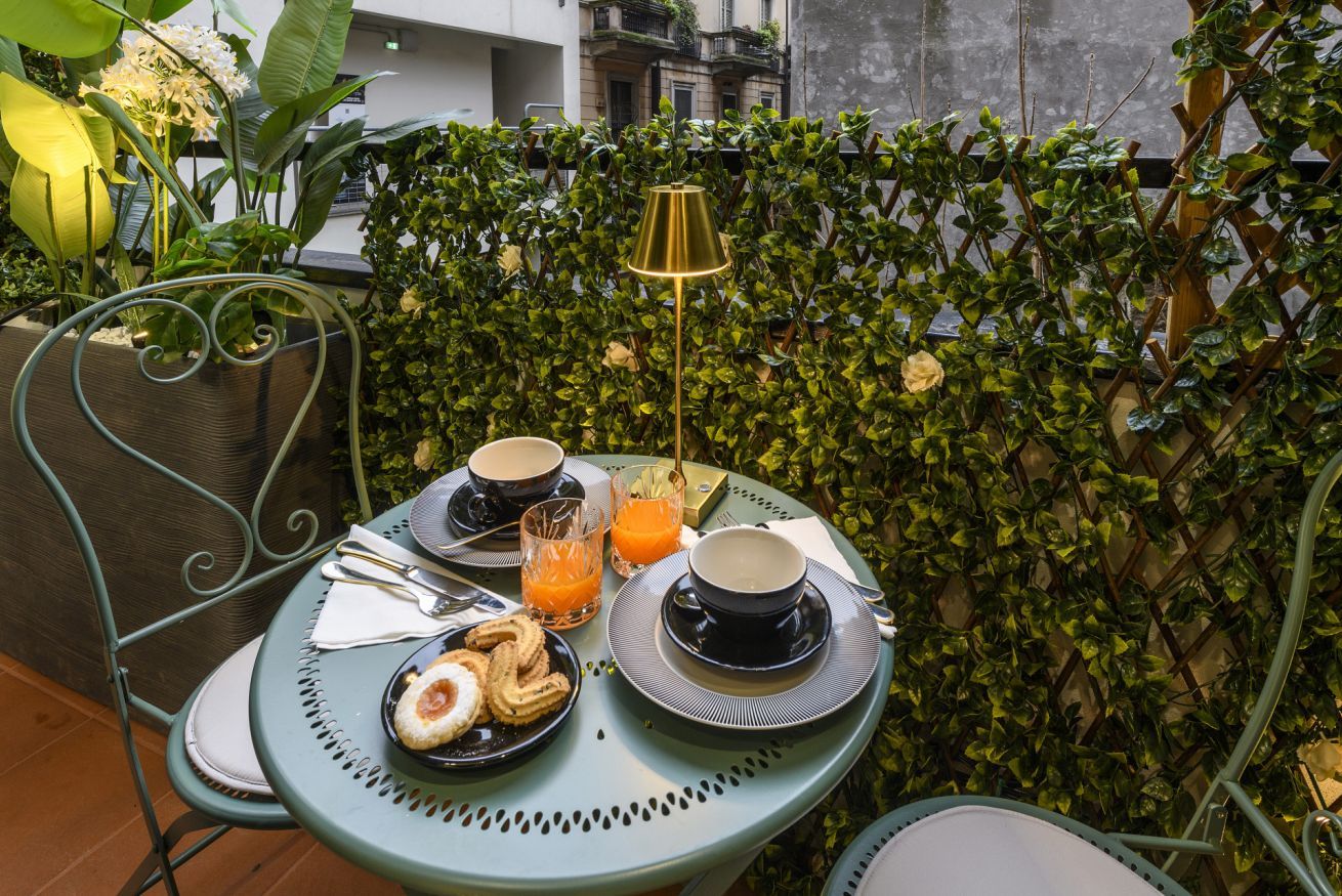 A table set with two cups, a glass of orange juice, and pastries, surrounded by greenery and a metal fence.