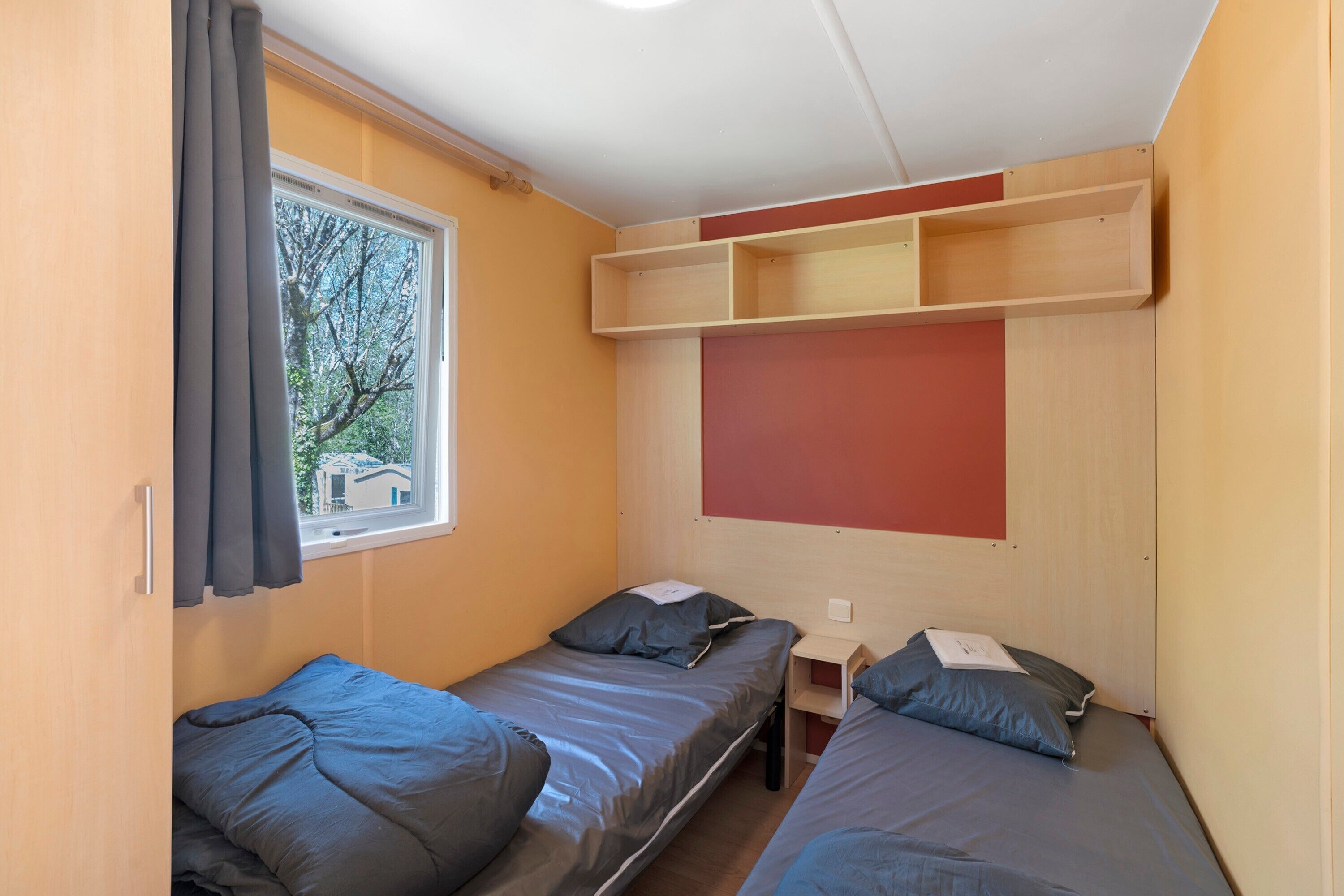 A small, compact room with two single beds, a window, and a built-in shelf above the beds.