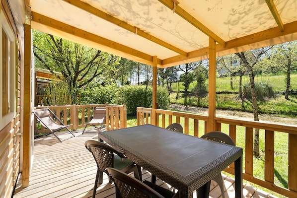 Une terrasse extérieure couverte, dotée d’une table et de chaises, et entourée d’une balustrade en bois et de verdure.