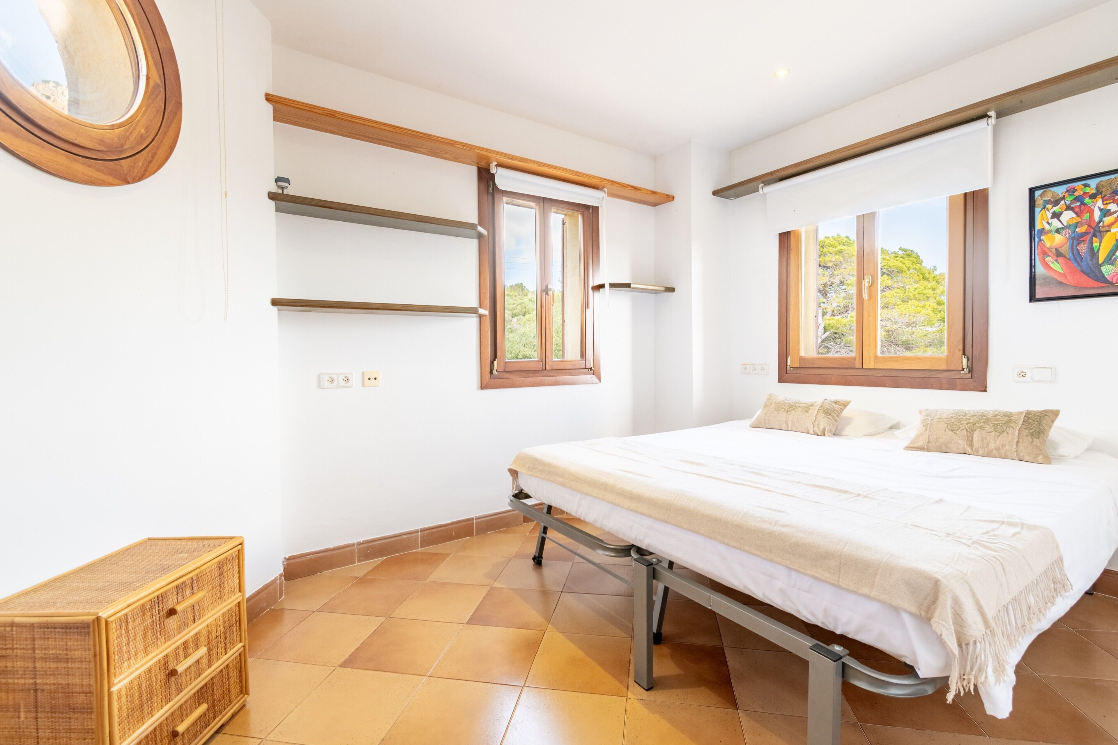 Un dormitorio con cama, ventanas, cabecera de madera, mesita de noche y estante fijado a la pared.