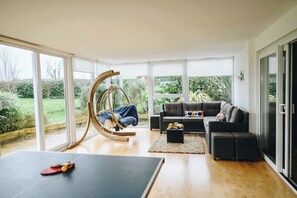 A spacious living room with a large sliding glass door leading to a garden, a rocking chair, a sofa with cushions, and a ping pong table. - Devon House Hot Tub Sea Views & Family Fun (Devon)