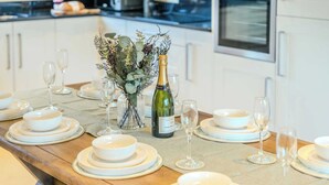 Ein Esstisch ist gedeckt mit weißen Tellern, Schüsseln und Weingläsern, dazu eine Champagnerflasche und ein Blumenstrauß.