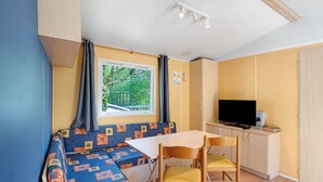 A compact room with a sofa, a dining table, a TV, and a window with curtains. - 3★ Campsite in Périgord Noir – Mobile Home with Pool, Fishing & Family Activities (Saint-Martial-de-Nabirat)