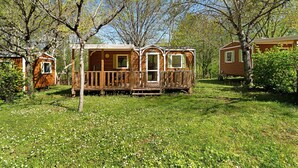 A wooden cabin with a deck, surrounded by trees and grass. - 3★ Campsite in Périgord Noir – Mobile Home with Pool, Fishing & Family Activities (Saint-Martial-de-Nabirat)