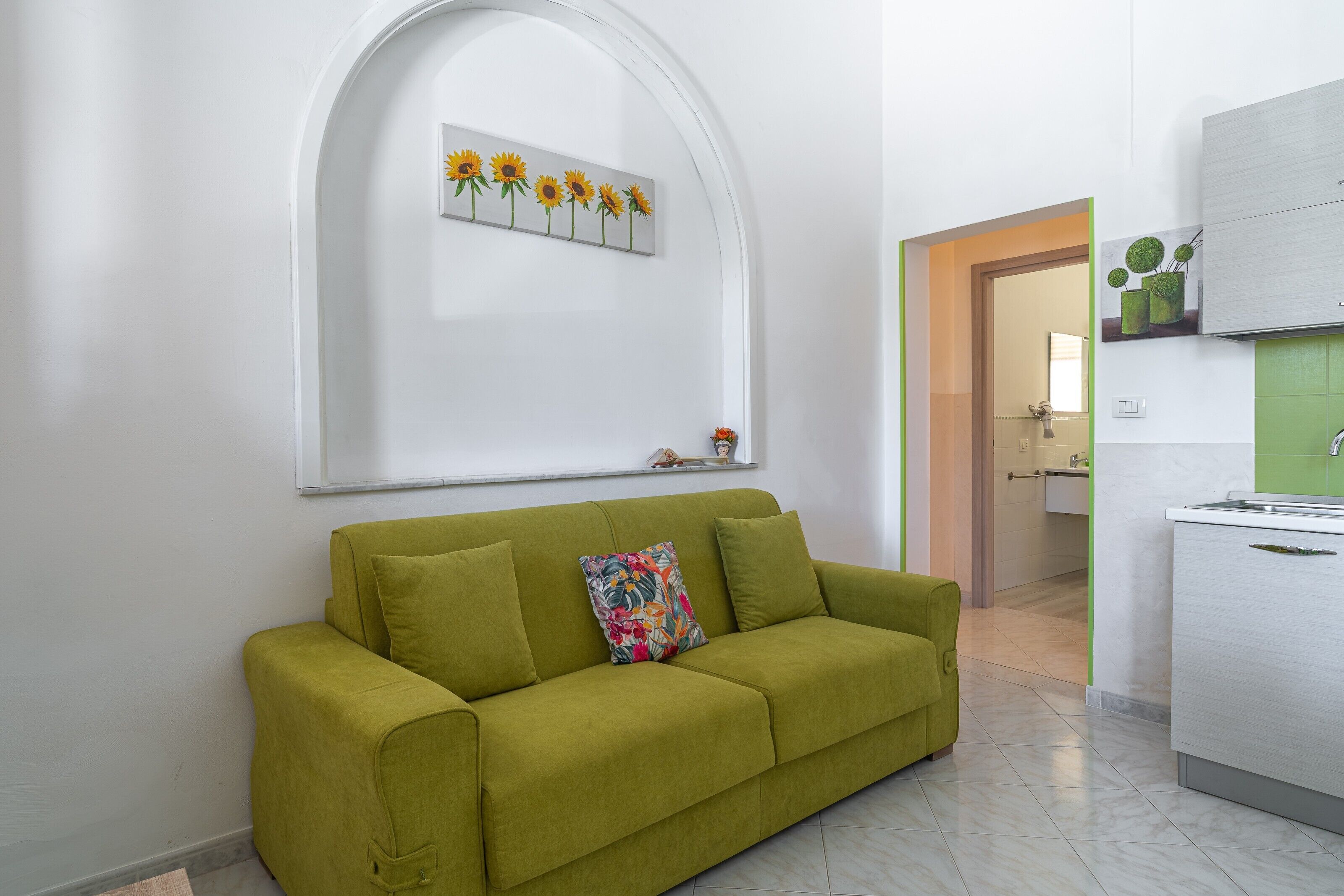 Ein grünes Sofa mit einem blumigen Kissen, einer an der Wand befestigten Sonnenblumen-Dekoration und ein Küchenbereich mit grüner Rückwand.