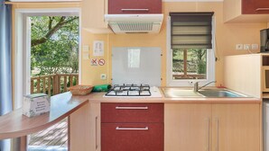 Une cuisine de taille réduite, équipée d’une cuisinière à gaz, d’un évier et d’une fenêtre avec des stores.