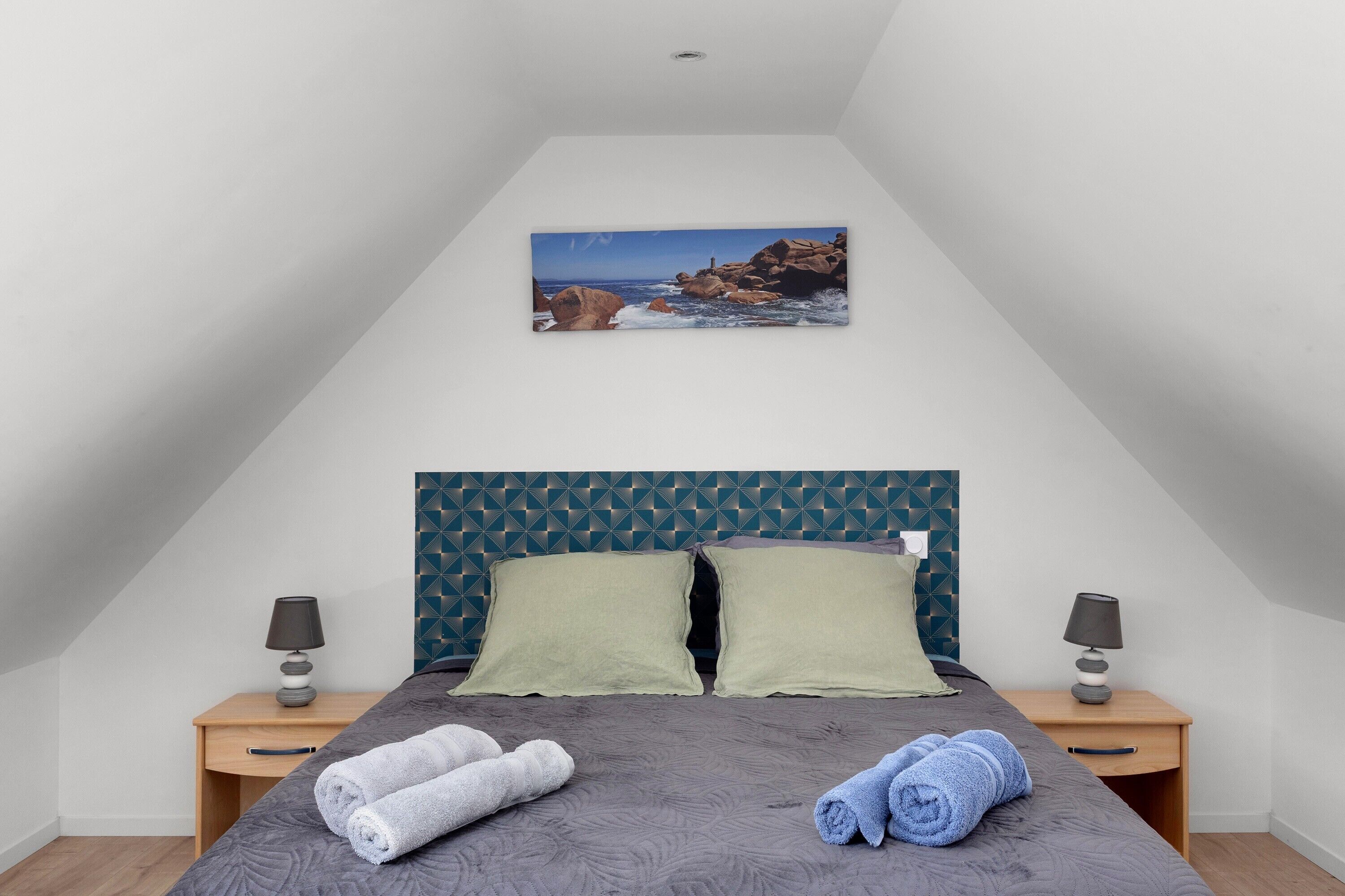 A bedroom with a bed, two pillows, two bedside tables with lamps, and a painting of a rocky coastline.