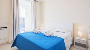 A hotel room with a large bed, a nightstand with a lamp, and a view of the city through the window. - Luxury Seafront Apartment Private Terrace Crescent (Salerno)