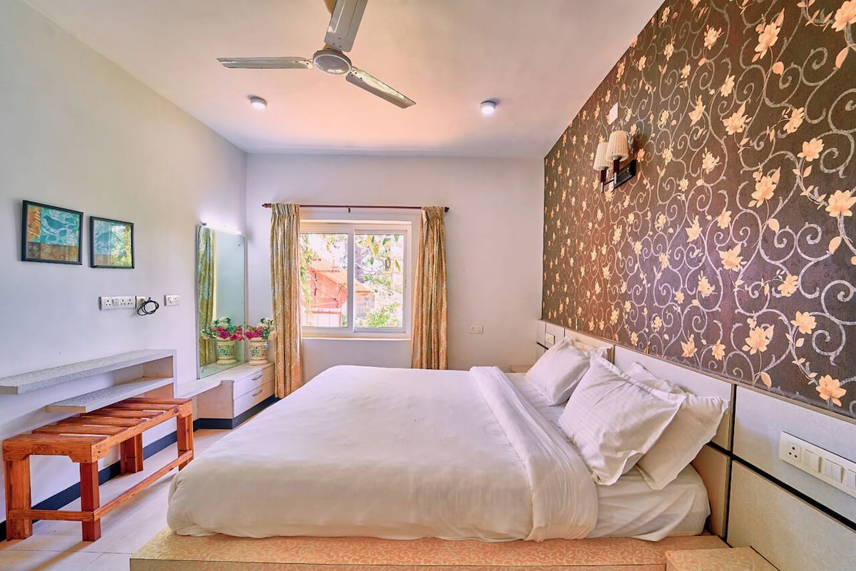 Un dormitorio con una cama, un ventilador de techo, una ventana con cortinas y una pared con estampado floral.
