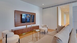 Ein modernes Wohnzimmer mit einem an einer Holzplatte montierten Flachbildfernseher, einem beigefarbenen Sofa, zwei weißen Sesseln, einem kleinen runden Couchtisch und einem Schrank unter dem Fernseher.