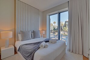 A modern hotel room with a large bed, two bedside lamps, a view of the outdoors, and a balcony with a chair. - NEW Beach Apartment in Quarteira (Loulé)