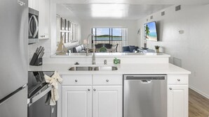 A modern kitchen with white cabinetry, stainless steel appliances, a large window with a view of the sea, and a dining area with a blue couch. - SUMMER DISCOUNT! Beachfront 2B/2B Vacation Condo Carlos Pointe 221. Amazing Views! EVERYTHING BRAND NEW! (Fort Myers Beach)