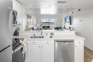 A modern kitchen with white cabinetry, stainless steel appliances, a large window with a view of the sea, and a dining area with a blue couch. - SUMMER DISCOUNT! Beachfront 2B/2B Vacation Condo Carlos Pointe 221. Amazing Views! EVERYTHING BRAND NEW! (Fort Myers Beach)