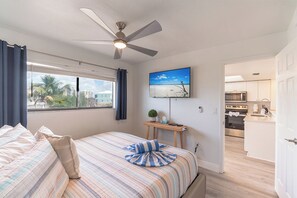 A bedroom with a bed, a ceiling fan, a TV, a small table with a plant, and a view of the kitchen. - SUMMER DISCOUNT! Beachfront 2B/2B Vacation Condo Carlos Pointe 221. Amazing Views! EVERYTHING BRAND NEW! (Fort Myers Beach)