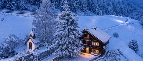 A snow-covered lodge with a large tree in front, surrounded by a forest.