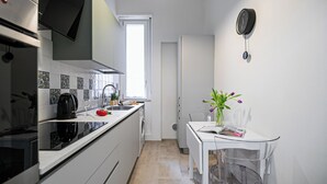 A compact kitchen with a built-in oven, a sink, and a window. There's a dining area with a table and chairs, and a vase with flowers on the table. - Italianway Comfort - Gomes 5 (Milan)