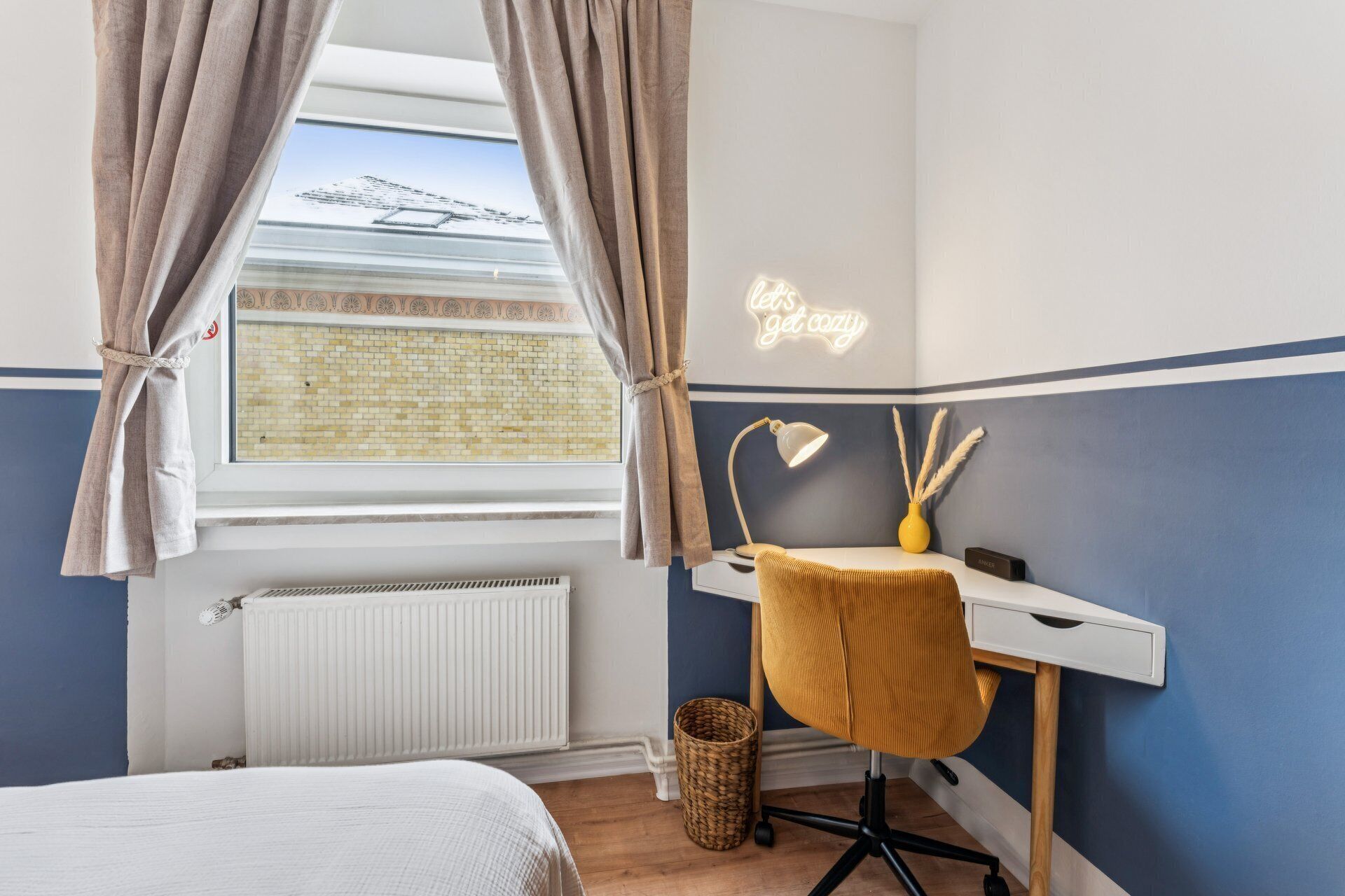 A bedroom with a bed, a desk with a chair, a radiator, and a window with curtains.