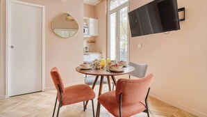 A dining area with a round table, two orange chairs, a TV mounted on the wall, and a kitchen in the background. - Confortable 3 pièces avec climatisation (Paris)