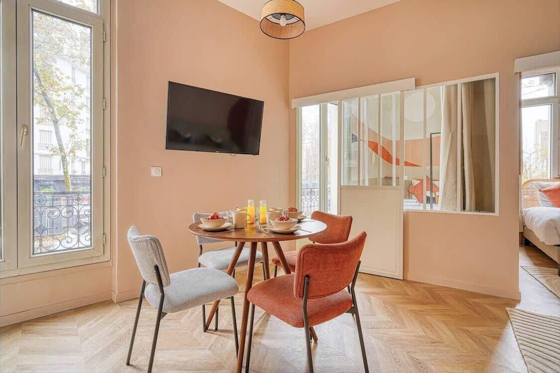A modern room with a dining table set for breakfast, a flat-screen TV mounted on the wall, and large windows offering a view of the outside.
