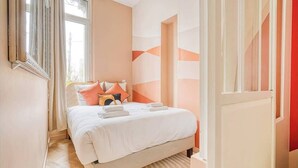 A neatly made bed with pillows and a blanket, a window with curtains, and a wall with abstract patterns. - Confortable 3 pièces avec climatisation (Paris)